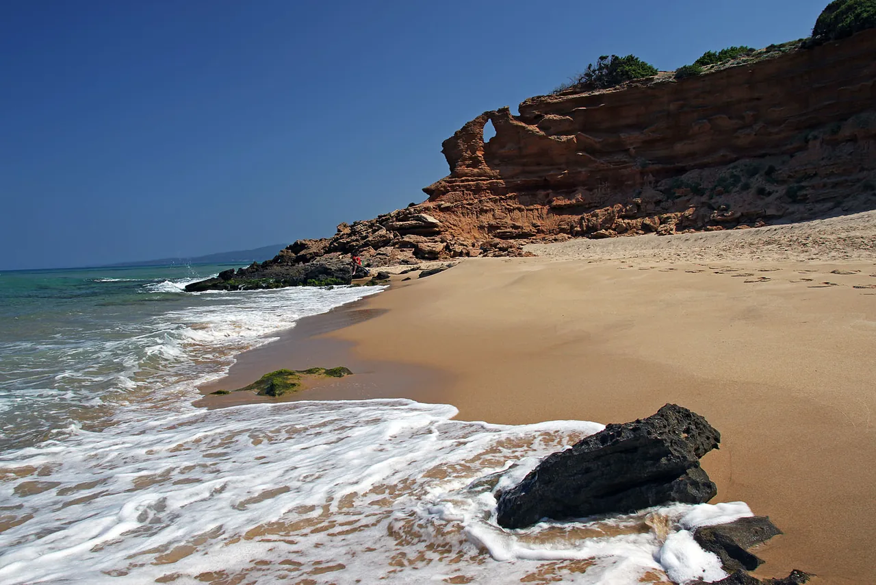 Wellen am Sandstrand von Scivu vor rötlichen Felsen.
